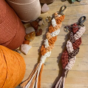 May include: Two handmade macrame keychains with tassels. One keychain is orange, white, and cream. The other keychain is brown, white, and cream. Both keychains have a silver key ring and a small charm.