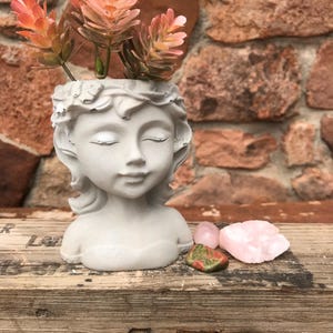 May include: A concrete planter shaped like a woman's bust with a crown of leaves. The planter is filled with pink and green succulents. Three pink and green stones are in front of the planter.