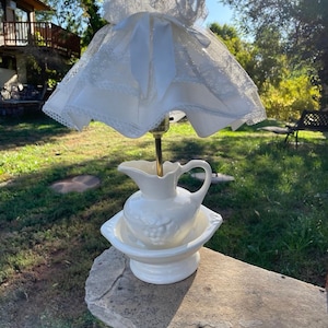 Puede incluir: Una lámpara vintage de cerámica blanca con base de jarra y cuenco, coronada por una pantalla de encaje blanco. La lámpara tiene un tallo de latón y está colocada al aire libre sobre una superficie de piedra, con césped verde y árboles.