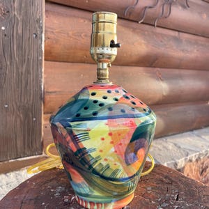 Puede incluir: Una colorida lámpara de mesa de cerámica con un casquillo de latón. La lámpara presenta un diseño abstracto vibrante con tonos verdes, amarillos, rojos y naranjas. Tiene una forma única y un cable amarillo.