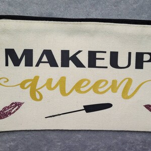 Può includere: Una trousse per il trucco in tela bianca con una cerniera nera. La trousse ha le parole "MAKEUP queen" stampate su di essa in lettere nere e dorate. Ci sono due piccole labbra di glitter rosa su entrambi i lati del testo.