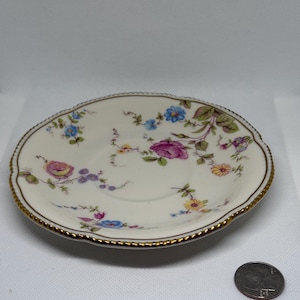 May include: A small, off-white saucer with a floral design in pink, blue, and yellow. The rim is scalloped and trimmed with gold. A US quarter is next to the saucer.