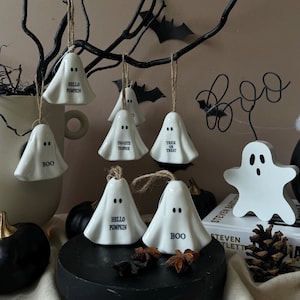 May include: White ceramic ghost ornaments with black eyes and text, including "BOO", "HELLO PUMPKIN", "TOO CUTE TO SPOOK", and "TRICK OR TREAT". The Halloween decorations are hanging from a black branch.