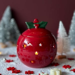 Op de afbeelding: Een rode keramische kerstbal wax warmer met een groen hulstontwerp en een witte wax smelt in de vorm van een sneeuwpop. De warmer heeft een lichtje erin en is omringd door witte sneeuw en rode sneeuwvlokken.