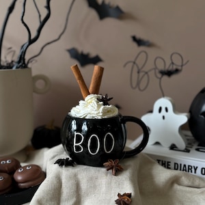 Könnte beinhalten: Schwarze Keramiktasse mit dem Wort "BOO" in weißen Buchstaben, gefüllt mit Schlagsahne, Zimtstangen und Sternanis. Die Tasse steht auf einer beigefarbenen Stoffoberfläche mit zusätzlichem Sternanis und Schokoladen-Marshmallows.