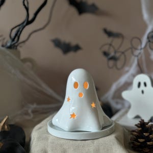 Może przedstawiać: Biały ceramiczny świecznik w kształcie ducha z wycięciami w kształcie gwiazd i okrągłą podstawą. Duch ma dwa otwory na oczy i świecące pomarańczowe światło w środku. Dekoracja na Halloween o upiornym motywie.