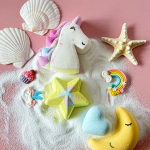 Puede incluir: Una colección de bombas de baño caprichosas y elementos decorativos sobre una superficie rosa. Incluye un unicornio, estrellas de mar, conchas marinas, una estrella, un corazón, una luna creciente, un arcoíris y una magdalena. Las bombas de baño son de varios colores.