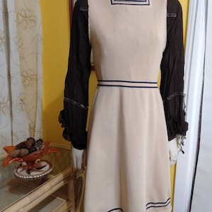 Puede incluir: Un vestido sin mangas de color crema con un ribete azul marino alrededor del escote y el dobladillo. El vestido está ajustado a la cintura y se ensancha hasta el dobladillo. El vestido se está modelando en un maniquí.