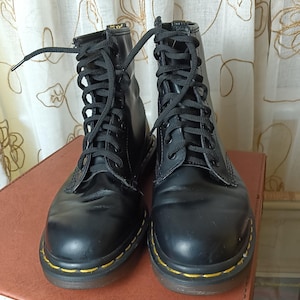 Puede incluir: Un par de botas de cuero negro con cordones, costuras amarillas y cordones negros.