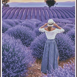 May include: A woman wearing a white shirt and a striped skirt stands in a field of lavender. She is wearing a white hat and is looking out at the lavender field. The field is in rows and there are trees in the background.