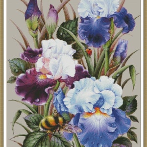 May include: A cross-stitch floral design featuring irises in shades of purple, white, and blue. A bumblebee and a ladybug are also present. The artwork is set against a neutral background and framed in a gold-colored border.