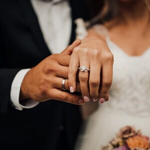 May include: A close-up of two hands with wedding rings. The woman's hand is on top of the man's hand, and she is wearing a diamond engagement ring.