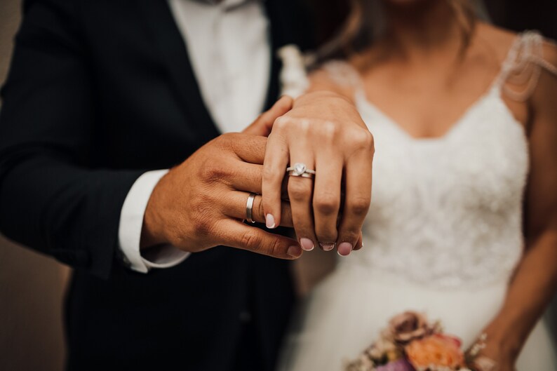 May include: A close-up of two hands with wedding rings. The woman's hand is on top of the man's hand, and she is wearing a diamond engagement ring.