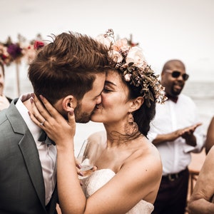 May include: A bride and groom kiss during their wedding ceremony on a beach. The bride is wearing a white dress and a floral crown. The groom is wearing a gray suit. Guests are in the background.