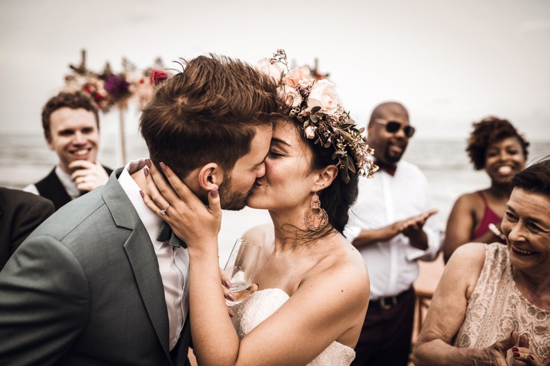 May include: A bride and groom kiss during their wedding ceremony on a beach. The bride is wearing a white dress and a floral crown. The groom is wearing a gray suit. Guests are in the background.