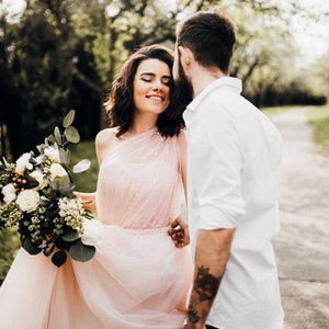May include: A couple in a forest setting. The woman is wearing a pink dress and holding a bouquet of white flowers. The man is wearing a white shirt and has a beard.