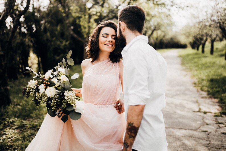 May include: A couple in a forest setting. The woman is wearing a pink dress and holding a bouquet of white flowers. The man is wearing a white shirt and has a beard.