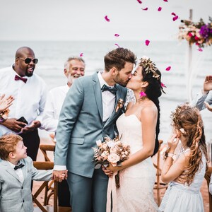 May include: A bride and groom kiss during their wedding ceremony on a beach. Guests are gathered around them, smiling and clapping. The bride is wearing a white strapless dress and a flower crown. The groom is wearing a gray suit and a white bow tie. Pink flower petals are falling from the sky.