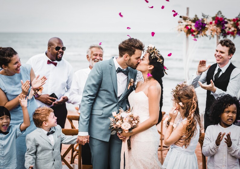 May include: A bride and groom kiss during their wedding ceremony on a beach. Guests are gathered around them, smiling and clapping. The bride is wearing a white strapless dress and a flower crown. The groom is wearing a gray suit and a white bow tie. Pink flower petals are falling from the sky.