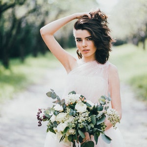 May include: A bride in a white dress with a one-shoulder neckline holds a bouquet of white flowers and greenery.