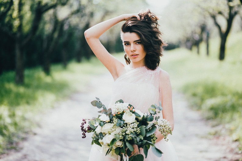 May include: A bride in a white dress with a one-shoulder neckline holds a bouquet of white flowers and greenery.