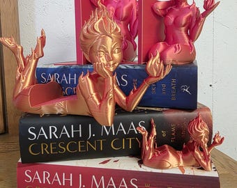 May include: A collection of 3D-printed mermaid figurines in shades of pink and gold, posed on top of books. The books are titled "Sarah J. Maas Crescent City" and "Sky and Breath". The figurines have flame-like hair and are holding flames.