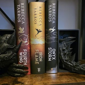 May include: Three books by Rebecca Yarros, "Fourth Wing", "Iron Flame", and "Onyx Storm", are displayed on a wooden shelf. The books are flanked by black dragon-themed bookends. The book spines are various colours, with gold and white lettering.