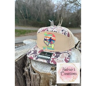 Könnte beinhalten: Beige und Camouflage Trucker-Cap mit farbenfrohem Aufdruck "Boats N' Hoes". Die Kappe hat einen Mesh-Rücken und einen gebogenen Schirm. Die Kappe liegt auf einem Baumstumpf.