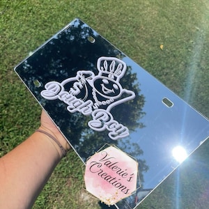 Puede incluir: Una placa de matrícula reflectante blanca con un gráfico blanco de un personaje de dibujos animados con una corona y el texto "Dough Boy" sobre un fondo negro. El texto "Valerie's Creations" es visible en el reflejo.