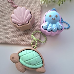 May include: Three small 3D printed keychains featuring a light pink seashell, a light blue octopus with white dots, and a light green turtle. Each figure is attached to a matching coloured ball chain, showcasing textured surfaces and distinct shapes.