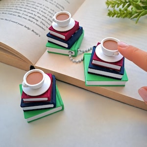 May include: Three miniature stacks of books, each topped with a small white teacup and saucer. The books are coloured in shades of green, dark blue, and maroon. A silver ball chain connects two of the stacks, which are arranged on an open book.