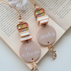May include: Two beige lanyards with book and bookshelf charms. Each lanyard has a round tag with a name printed on it. One says "Miss McPherson" and the other says "Sandra". Both have gold-coloured clasps.