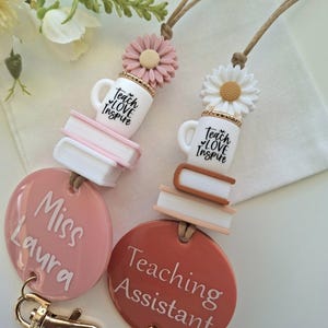 May include: Two lanyards featuring silicone beads shaped like daisies, coffee mugs with the text Teach LOVE Inspire, and stacked books. One lanyard has a pink circular tag with Miss Laura, the other a terracotta tag with Teaching Assistant.