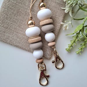 May include: Two beaded lanyards with gold-coloured clasps. The beads are white, grey, and natural wood, with gold accents. The lanyards are displayed on a tan fabric background.