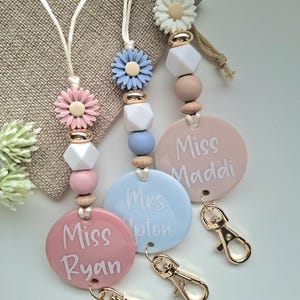 May include: Three teacher lanyards with daisy and geometric bead accents. Each lanyard features a round, pastel-coloured name tag with a teacher's name in white script. Gold-coloured lobster clasps are attached.