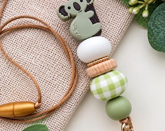 May include: A lanyard featuring a silicone koala charm, a white bead, a small gold-toned spacer, a natural wood-toned bead, a green and white gingham patterned bead, and a solid sage green bead, attached to a gold-toned metal lobster clasp.