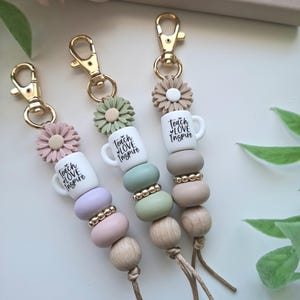 May include: Three keychains featuring gold-tone lobster clasps, silicone daisy beads, and white mug-shaped beads printed with the text "teach LOVE Inspire." Each includes a mix of pastel silicone, metallic gold, and natural wood beads with cord ends.