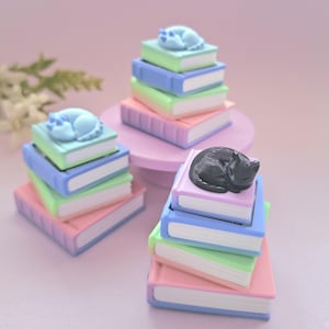 May include: Three stacks of miniature books in pastel shades of pink, green, blue, and purple. Each stack is topped with a small figurine of a sleeping animal, with two light blue figures and one black figure resting on the top book covers.