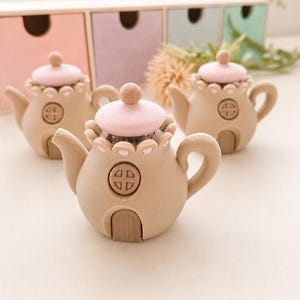 May include: Three beige teapot-shaped ornaments with pink lids and small round knobs. Each teapot has a small door and a circular design on the front. The teapots are arranged on a white surface.