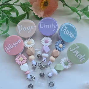 May include: Five circular badge reels in pastel colours, each personalised with a white name: Allison, Page, Emily, Olivia, and Jade. Each reel features a decorative silicone daisy flower, geometric beads, and a clear plastic strap with a metal snap.