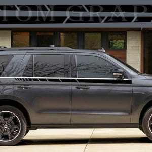 Op de afbeelding: Een grijze Ford Expedition SUV met zwarte zijstrepen. Het voertuig staat geparkeerd voor een huis met een houten deur.