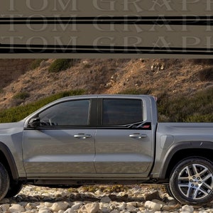 Puede incluir: Una camioneta plateada con detalles negros y calcomanías "ZR2" en el lateral. La camioneta está estacionada en una playa rocosa.