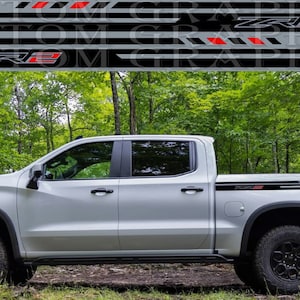 May include: A white Chevrolet Silverado pickup truck with black and red decals on the sides. The truck is parked in a wooded area.