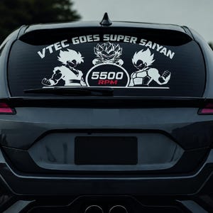 Op de afbeelding: Een zwarte auto met een sticker op de achterruit. De sticker heeft witte cartoonfiguren en de tekst "VTEC GOES SUPER SAIYAN" erboven. Daaronder staat "5500 RPM" in een rode cirkel. De auto staat op een weg.