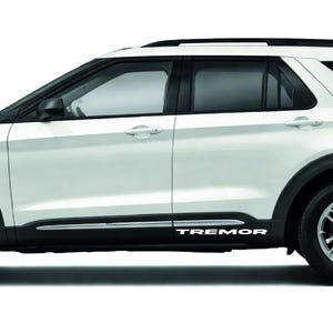 Op de afbeelding: Witte SUV met zwarte accenten, waaronder de wielkasten, het dak en de onderste bekleding. Het woord "TREMOR" staat in witte letters op de zijkant. Zilverkleurige lichtmetalen velgen en getinte ramen.