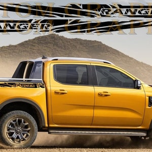 Könnte beinhalten: Ein gelber Ford Ranger Pickup mit schwarzen und weißen Grafikaufklebern an den Seiten. Die Aufkleber zeigen das Wort "Ranger" und abstrakte Designs. Der LKW steht auf einem Feldweg mit Hügeln im Hintergrund.