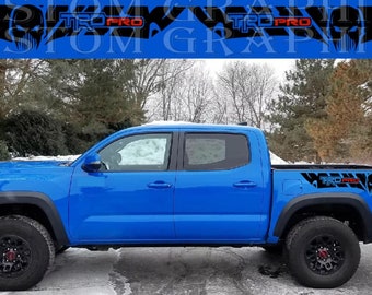 Könnte beinhalten: Ein blauer Toyota Tacoma Pickup mit schwarzen und roten "TRD PRO" Aufklebern an den Seiten. Der Pickup hat schwarze Räder und eine schwarze Ladefläche.