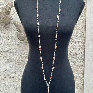 Può includere: Una lunga collana di perline bianche con accenti di conchiglie marroni e bianche. La collana ha un grande ciondolo a conchiglia nella parte inferiore.