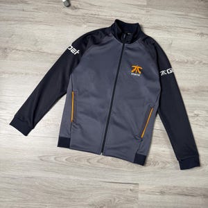 May include: A charcoal gray and black zip-up track jacket featuring a stand-up collar, long sleeves with ribbed cuffs, and two side pockets with orange trim. The Fnatic logo is printed in orange on the chest, with white text on the sleeves.