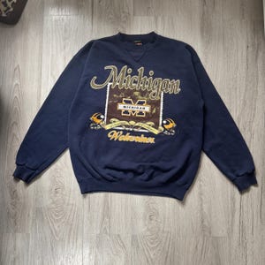 May include: A navy blue long-sleeve sweatshirt featuring a graphic print with the text Michigan, Wolverines, and A Tradition of Excellence. The design includes a central block with a yellow M logo, two football helmets, and decorative scrollwork.